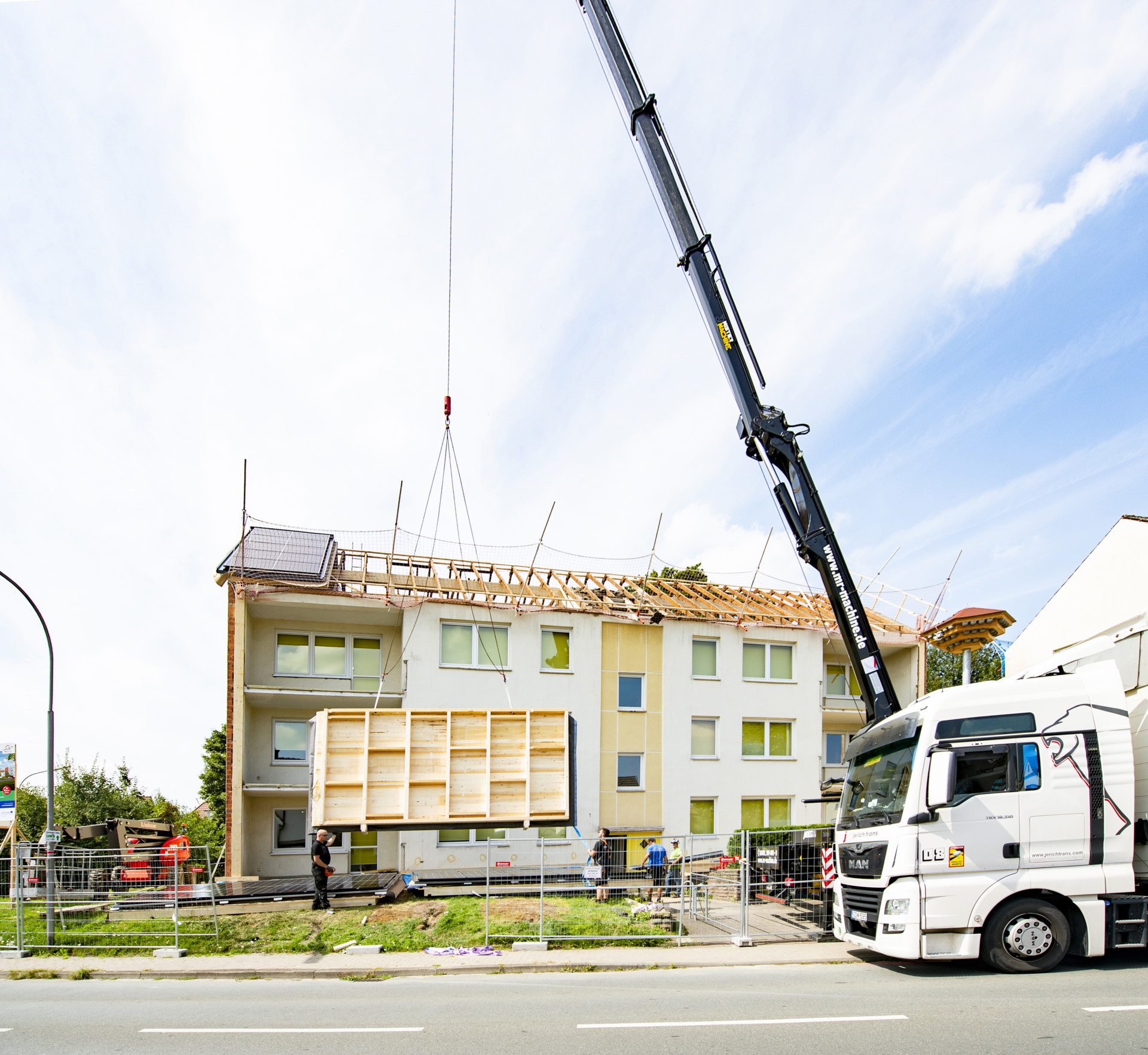 Foto, Blick von der Straße auf ein mehrstöckiges Wohngebäude. Das Dach liegt offen, ein Kran hebt ein vorgefertigtes Dachmodul in die Höhe.