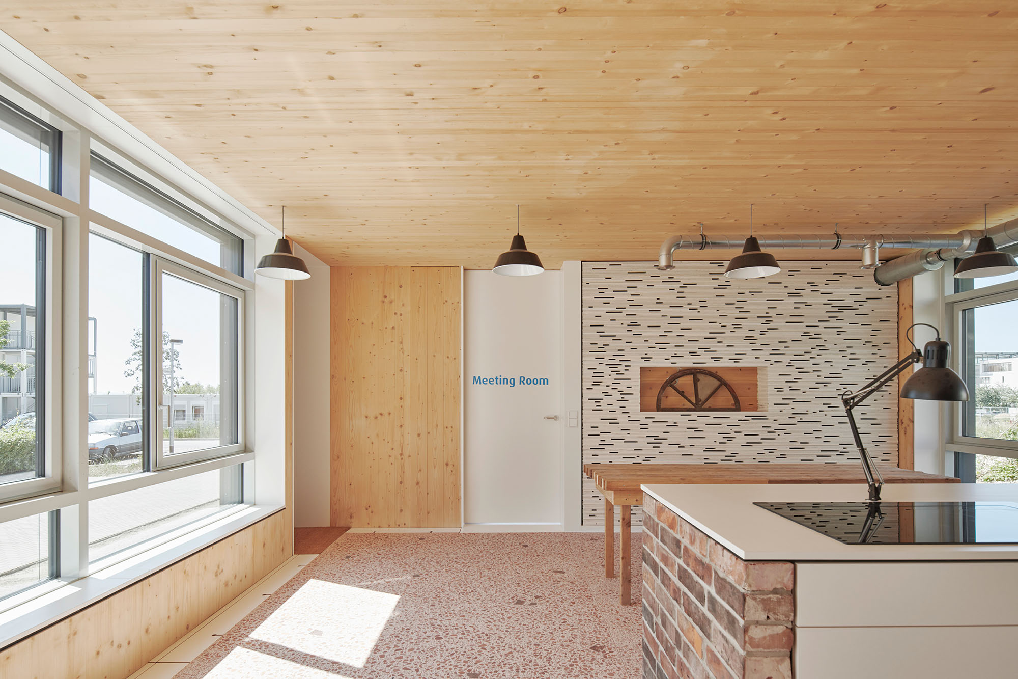 Foto, Innenraum mit Fenstern auf der linken Seite, Kücheninsel und große, Tisch links. Es sind verschiedene Materialen wie Backstein, Holz und Mosaik verbaut. 